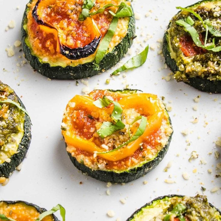 Close up of a few sliced zucchini pizza bites on a white tray. One has red sauce, a sliced yellow pepper and some fesh basil, another has pesto and roasted red pepper.