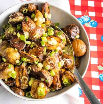 White bowl filled with grilled potatoes with mustard sauce and scallions on a red checkered background
