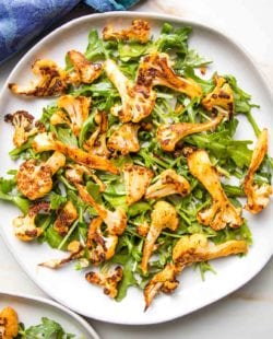 Close up of a white plate, with arugula and roasted cauliflower with paprika. The edge of a blue napkin in the upper left corner.