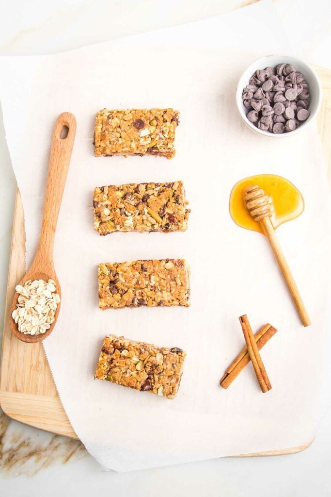 4 crunchy granola bars lined up on a piece of parchments. There is a spoon full of oats, a puddle of honey, a dish of chocolate chips and two cinnamon sticks.