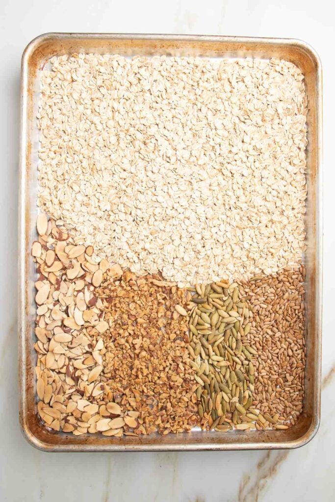 Large rimmed baking tray with oats, sliced almonds, chopped walnuts, sesame seeds and pumpkin seeds