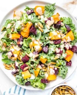 Large rimmed white plate filled with kale, roasted butternut squash and beets, toasted pumpkin seeds and a creamy balsamic dressing on top of a white tea towel with blue stripes and a small dish of extra toasted pumpkin seeds.