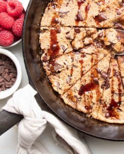 Cast iron skillet with a natural linen napkin tied around the handle, filled with chocolate chunk skillet cookies drizzled with date syrup and a spoon in the center pulling out an ooey, gooey piece. A small white dish of chocolate chunks and a dish of bright red raspberries are also in the photo.