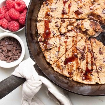 Cast iron skillet with a natural linen napkin tied around the handle, filled with chocolate chunk skillet cookies drizzled with date syrup and a spoon in the center pulling out an ooey, gooey piece. A small white dish of chocolate chunks and a dish of bright red raspberries are also in the photo.