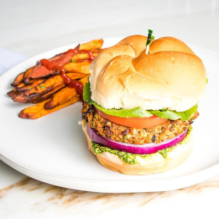 Veggie burger on a bun with green arugula aioli, purple onion, lettuce and tomato on a bun with sweet potato fries on the side of a round white rimmed plate.