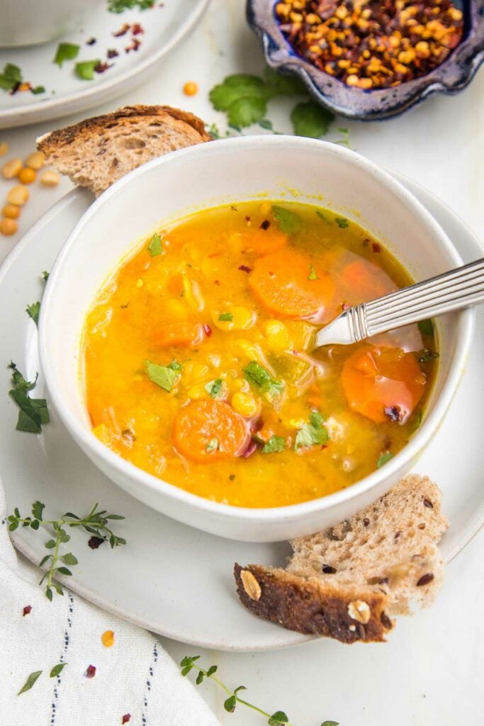 Bowl of yellow split pea soup with chunky carrots and some crusty bread. Fresh herbs and crushed red pepper sprinkled about.