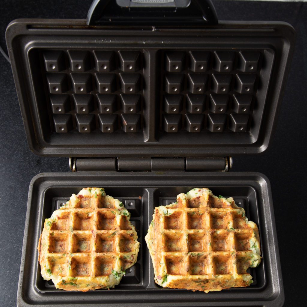 Finished waffles in iron with large specks of green herbs.