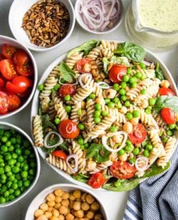 A white bowl filled with pasta salad and small bowls of the ingredients surrounding it: chick peas, green peas, halved cherry tomatoes, sunflower seeds, shallots and tahini parsley dressing.