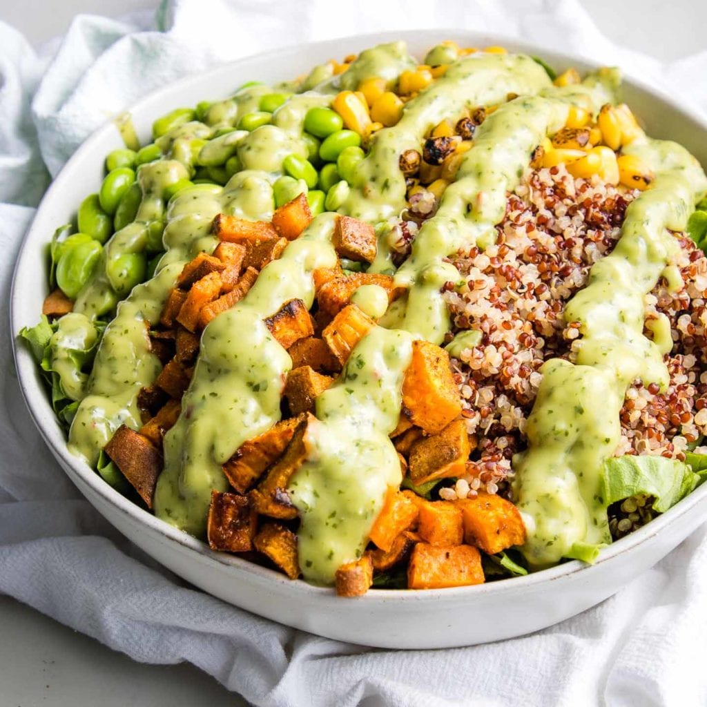 White bowl filled with greens, roasted sweet potatoes, quinoa, edamame beans and corn, drizzled with avocado lime cilantro dressing