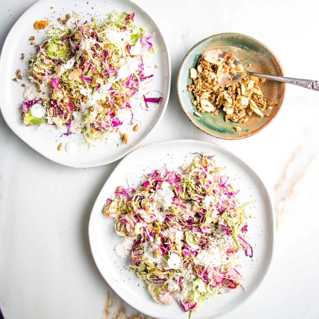 Brussels Sprouts Salad on two white plates with colorful red cabbage, drizzled with a creamy, dairy-free dressing and some toasted nuts/seeds with a little bowl of nuts and seeds and a spoon.