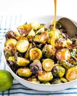 A stream of sweet and spicy sauce being poured onto a white bowl filled with crispy oven roasted brussels sprouts. There's a lime in the foreground and a teal striped towel underneath the bowl.