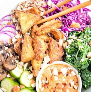 Close up of a brightly filled bowl of vegan food. Crispy triangles of tofu, sliced purple radishes, bright garlicky greens, sauteed mushrooms, tri-color quinoa, and a small bowl of almond butter dressing. Chopped almonds scattered over everything.