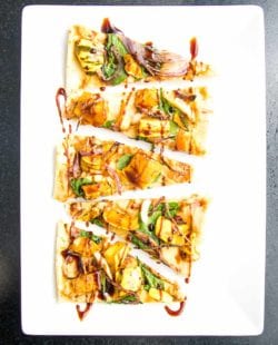 White plate with appetizer sized pieces of vegan flatbread topped with hummus, spinach and roasted squash, onions and garlic. Drizzled with date syrup.