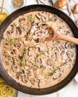 Cast iron skillet filled with sliced mushrooms in a creamy sauce. Around the skillet are some raw mushrooms, a small dish of pasta, a little glass dish with nutritional yeast and one with ground black pepper.