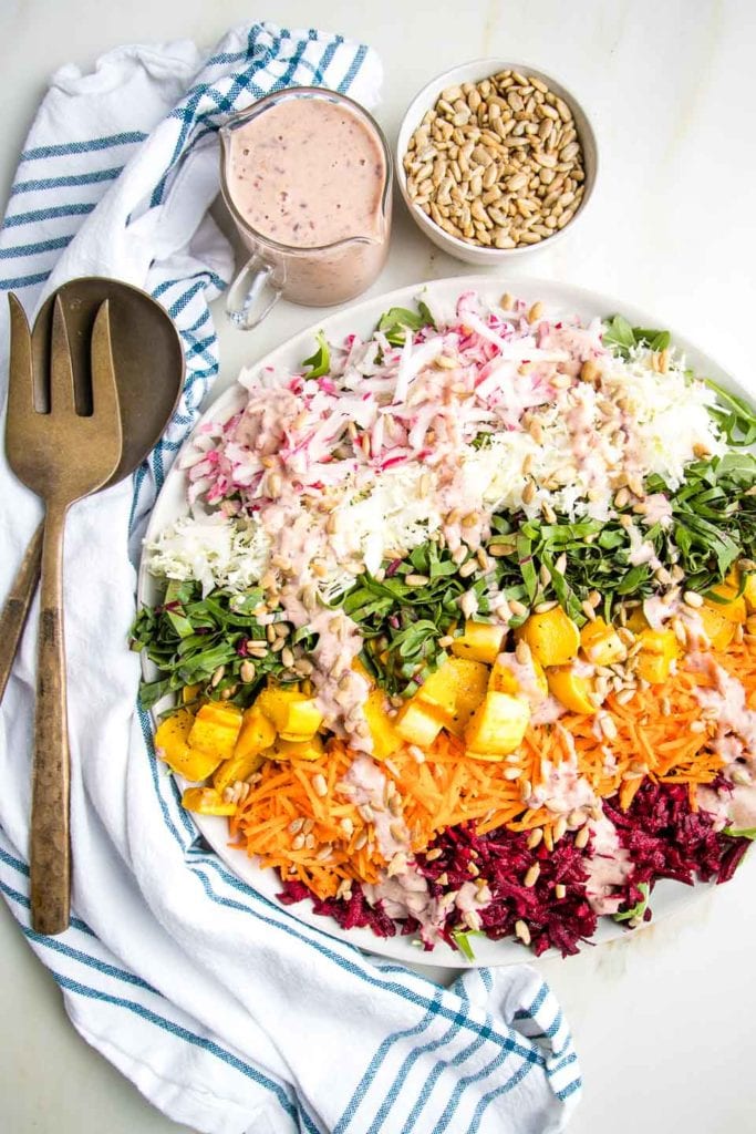 Shredded Rainbow Salad: radishes, cabbage, beet greens, carrots, red beets and roasted delicata squash cubes on a white rimmed plate, plus a small cut of pink dressing and a bowl of sunflower seeds.