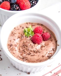 Super rich chocolate pudding in a small white dish, sprinkled with dark chocolate shavings, a sprig of fresh mint and 3 raspberries, with a small dish of assorted berries in the background.