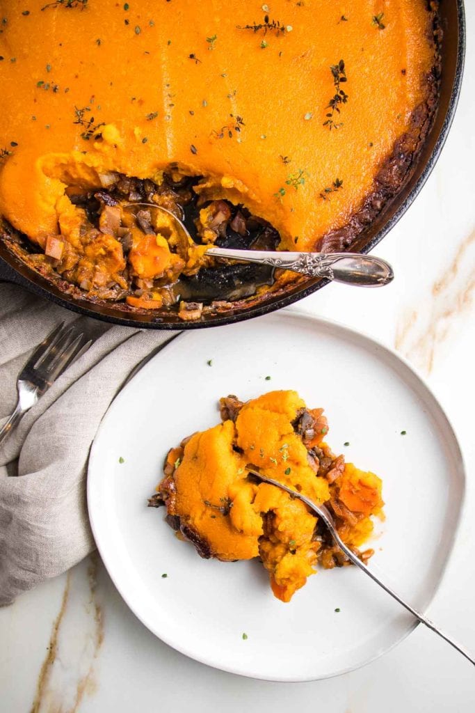 Cast iron skillet with cooked veggie shepherd's pie topped with sweet potato mash. A white rimmed plate is filled with a piece of shepherd's pie and a fork cutting through the middle. of it.