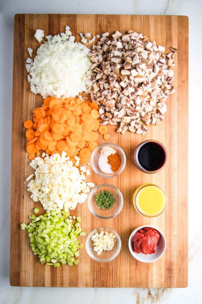 Large cutting board with ingredients for vegan shepherd's pie: chopped onions, carrots, parsnips, celery, mushrooms. Red wine, veggie broth, tomato paste, spices, fresh thyme and minced garlic.