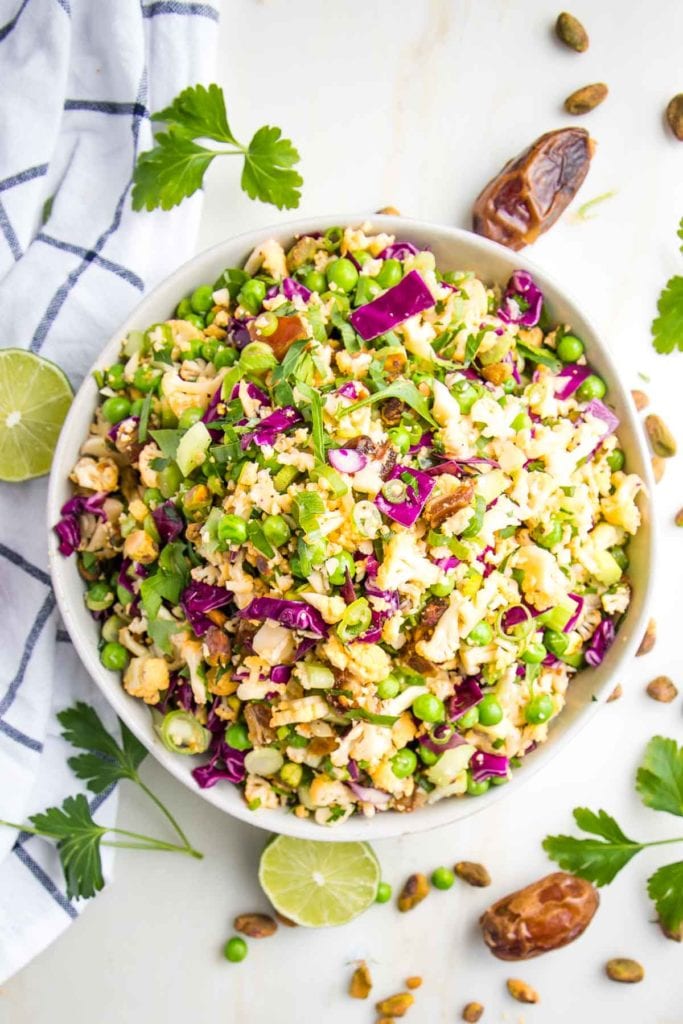 Large white bowl, filled with chopped colorful salad that includes purple cabbage, scallions, shaved cauliflower, fresh parsley, sliced scallions, green peas, diced dates. There are fresh limes, dates, fresh parsley and a dish towel strewn around the salad bowl.