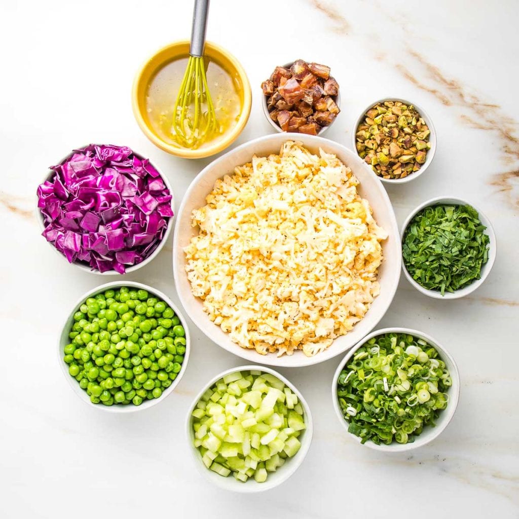 Bowls of ingredients for raw cauliflower salad. chopped cabbage, green peas, diced celery, sliced scallions, chopped parsley, pistachios, diced dates and a lime vinaigrette. All small bowls positioned in a circle, surrounding a white bowl of shaved cauliflower.