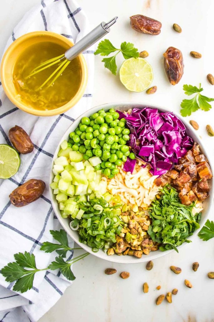 small yellow bowl of salad dressing and a large bowl of chopped colorful veggies each in their own section: purple cabbage, green peas, celery, chopped fresh parsley, pistachios, diced dates, and super thinly sliced cauliflower. Strewn around on the counter are pistachios, fresh parsley sprigs, dates, fresh limes and a dish towel.