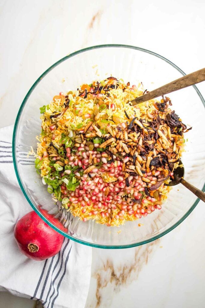 Large glass bowl filled with yellow rice, sliced scallions, pomegranate seeds, golden raisins and caramelized onions.