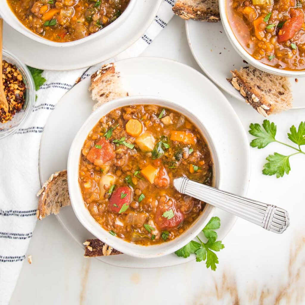 chunky vegetable lentil soup with colorful carrots, parsnips, tomatoes and parsley in a white bowl on a white plate. There are hand torn bits of crusty bread and fresh parsley strewn about.