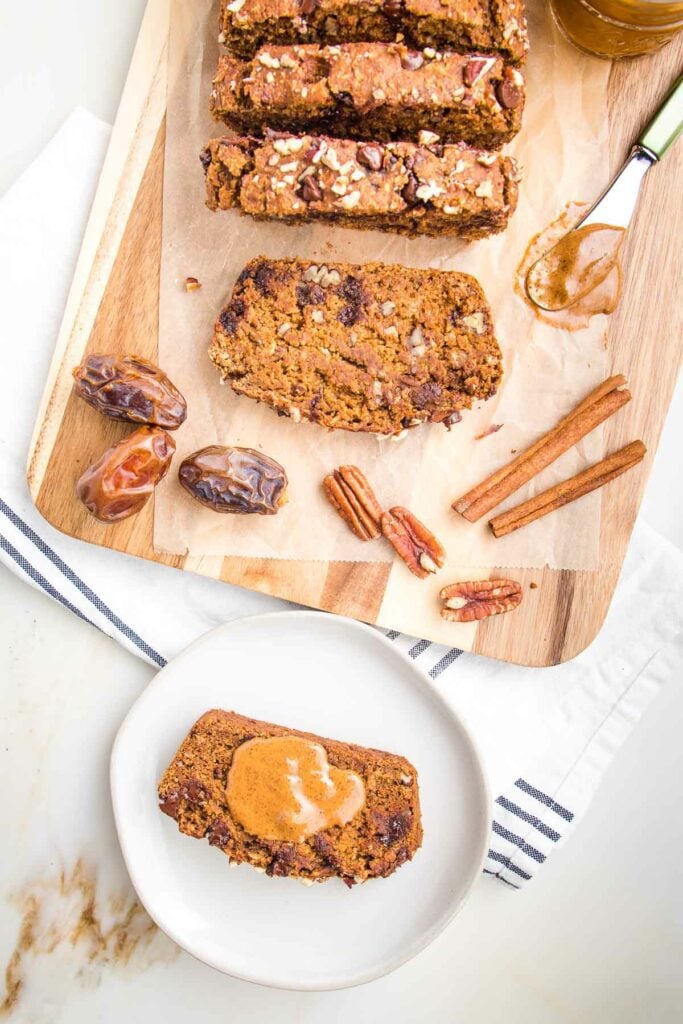 Slices of pumpkin bread with chocolate chips and pecans on a piece of parchment on a cutting board. In the foreground there is a white rimmed plate with a slice of pumpkin bread and a dollop of almond butter on top.