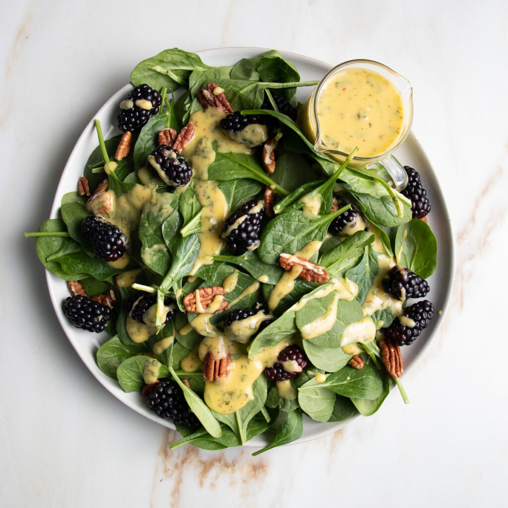 Spinach salad with blackberries, pecans and drizzled with mango dressing. There is a small glass pitcher with extra yellow, mango dressing on the side of the round white rimmed plate.