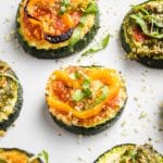Close up of a few sliced zucchini pizza bites on a white tray. One has red sauce, a sliced yellow pepper and some fesh basil, another has pesto and roasted red pepper.