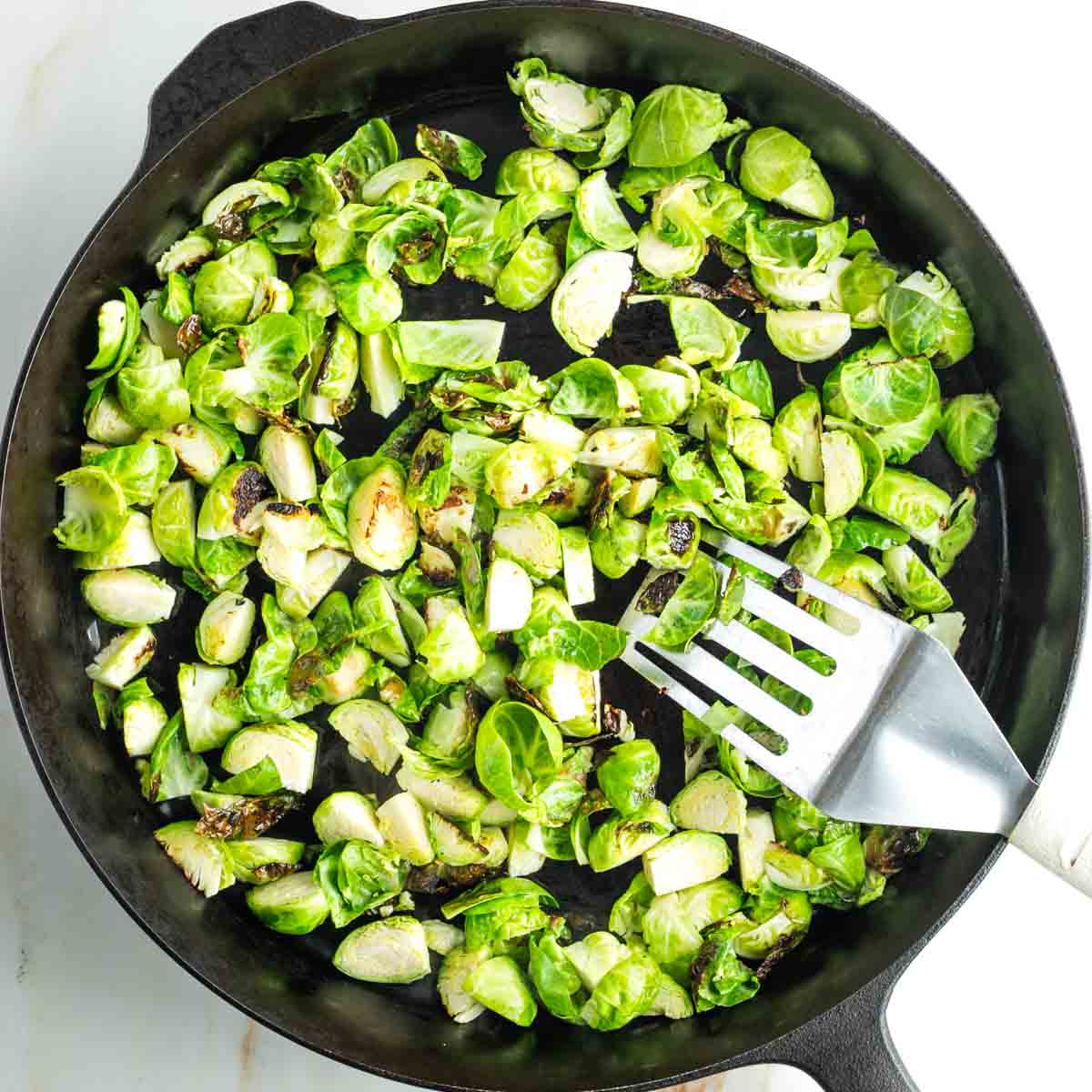Spatula pushing around quartered Brussels sprouts in a cast iron skillet as they start to get crispy. 