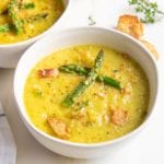 White bowl with yellowish-green thick soup, roasted asparagus tips floating in the center, and there is cracked black pepper and croutons sprinkled around. You can see another bowl of the same off to the upper left.