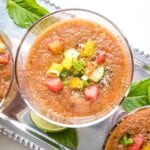 Close up of a glass filled with watermelon gazpacho with small diced, colorful veggies and cracked black pepper. Glass is on a silver tray with other glasses of gazpacho and also some mint leaves and slices of lime.