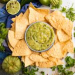 Green tomatillo salsa verde in a small bowl, surrounded by tortilla chips with a lime, a fresh tomatillo and some cilantro surrounding.