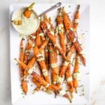 White platter with whole roasted and coated with spices carrots. A small glass bowl of dipping sauce with a few carrots in there and a spoon. The sauce is greenish and it's also drizzled on the cooked carrots. There are fresh herbs scattered around.