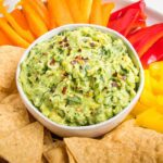 Close up of a bowl of chunky vegan guacamole sprinkled with red pepper flakes. The bowl is surrounded by tortilla chips and assorted color strips of bell peppers.
