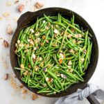 Cast iron pan filled with green beans almondine. There are almond slices, cloves of garlic and crushed red pepper scattered around the pan and a grey napkin tied around the cast iron handle.