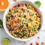 close up photo of a large white bowl filled with colorful quinoa salad that includes red peppers, green edamame, black beans, bright yellow corn and chopped cilantro. There is an orange half and a half a lime, plus fresh cilantro leaves around the bowl.