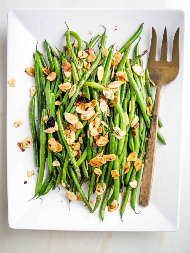 White rectangular dish with Green beans almondine. Bright green string beans loaded with sliced almonds and crushed red pepper with a bronze colored serving fork.