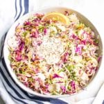Large white bowl with brussels sprouts slaw: colorful red cabbage, shredded brussels sprouts, various nuts and seeds, lemon wedge and a creamy vegan dressing.