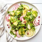Large rimmed white plate with lemony arugula salad. Thinly sliced radish, quartered artichoke hearts, thinly sliced avocado plus pomegranate seeds and lemon slices decorate the simple salad.