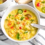 white bowl, filled with corn chowder, topped with colorful chunks of vegetables. There is a spoon I the bowl and the edges of other bowls of soup can be seen. The chowder is on top of a white linen napkin with some dark blue thin stripes.