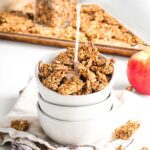 Pouring milk onto a bowl of granola with a tray of granola in the background and crumbs all around, plus a red apple.