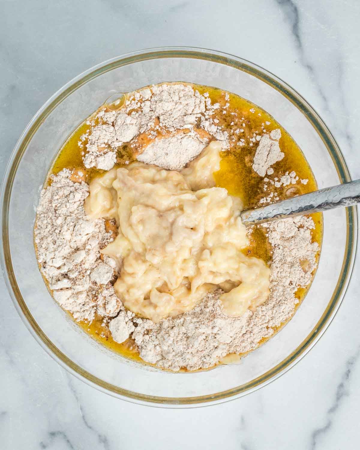 Wet ingredients, dry ingredients and mashed bananas in a bowl mixing up a banana bread. 
