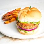 Veggie burger on a bun with green arugula aioli, purple onion, lettuce and tomato on a bun with sweet potato fries on the side of a round white rimmed plate.