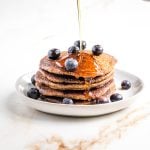 Syrup pouring onto stack of pancakes with blueberries on top.