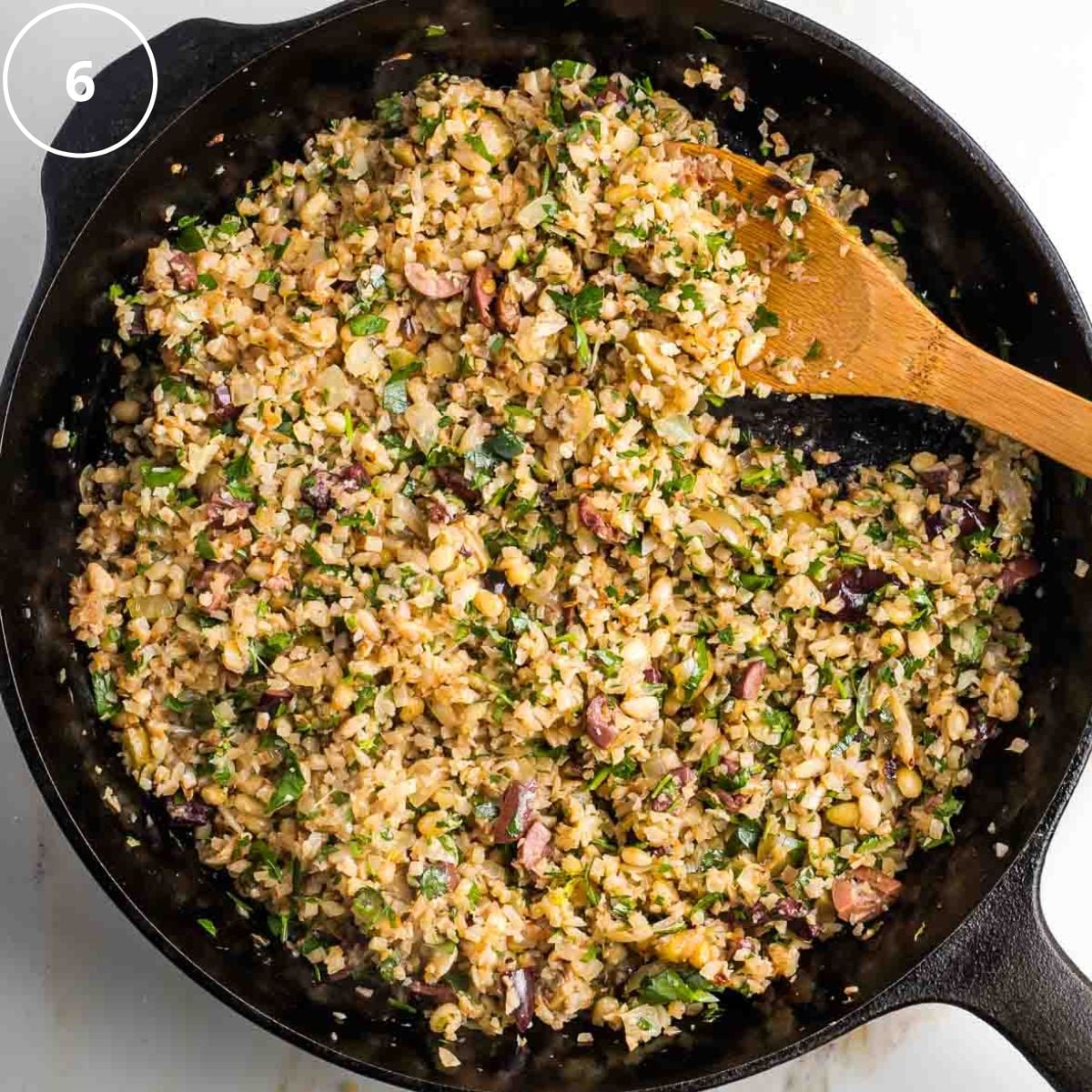Wooden spoon stirring mediterranean cauliflower rice in a cast iron skillet with olives, fresh herbs and pine nuts.