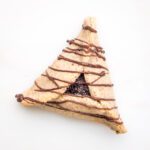 Poppy seed filled hamantaschen with chocolate drizzles across it, on a white background.