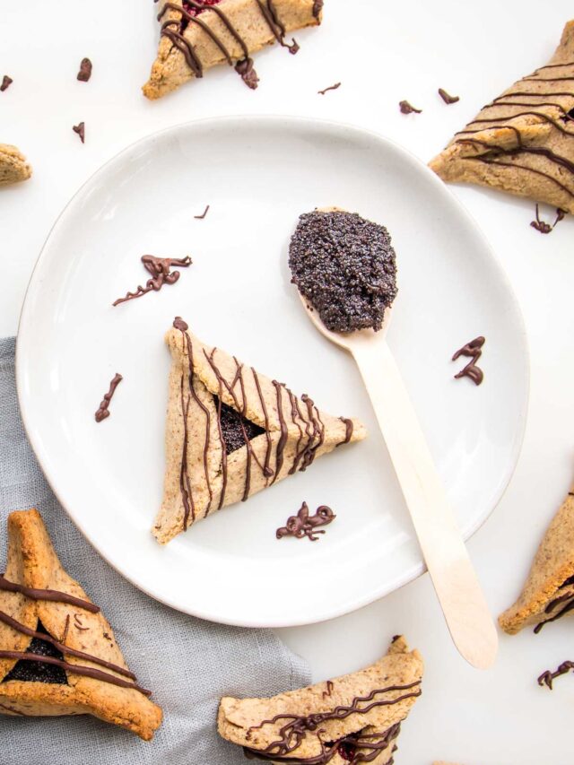 plate of hamentaschen with poppy seed filling and a spoonful of extra filling. Chocolate is drizzled on hamentaschen. There are more cookies scattered around.