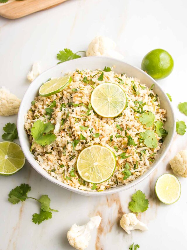 A large white, shallow bowl, filled with cauli-rice with limes and fresh cilantro. A sprinkling of ingredients around the bowls edge.